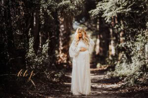 Pregnant woman standing on forest path in flowing white lace maternity gown, moody green maternity session location with soft directional light